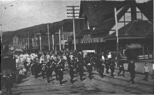 Black-and-white street scene of the Shamokin Band marching past the Reading Station with onlookers nearby