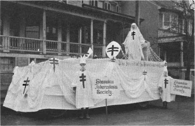 Black-and-white photograph of a decorated tuberculosis-committee parade float with double-barred cross emblems and a costumed figure